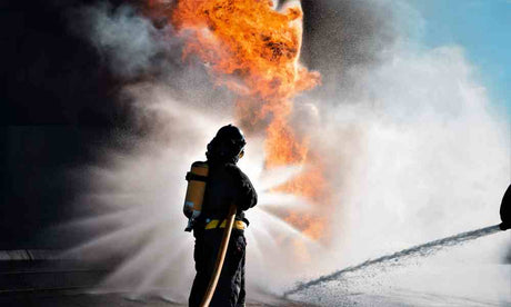 A wildland firefighter in full gear with a helmet is seen from behind as they spray a huge blaze with a firehose.