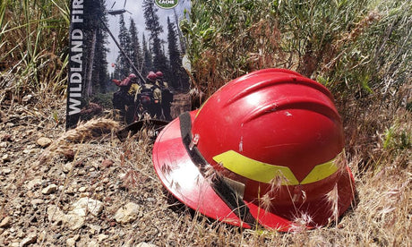 A red wildland firefighter helmet with yellow reflective strips rests on rocky ground beside a photo of firefighters.