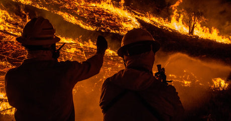 Two wildland firefighters watch a large wildfire while one holds a fire hose and the other communicates on a walkie-talkie.