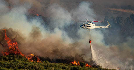 A white helicopter hovering over a large forest fire while using a bambi bucket to stop the fire from spreading.