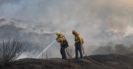 Two wildland firefighters hold a water hose pointed down the edge of a cliff. Smoke billows in the distance.