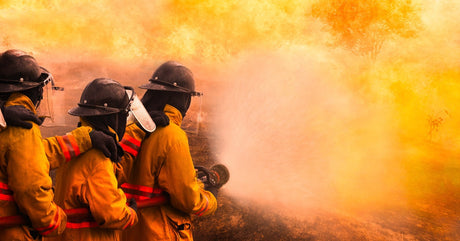  Three wildland firefighters wearing protective equipment stand in a line while holding a water hose toward fire.