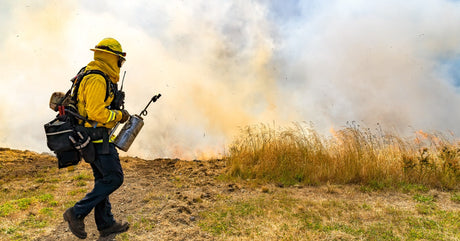 A wildland firefighter wearing yellow protective gear and goggles holds equipment while walking near a grass fire.
