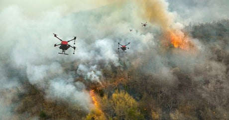 A group of four black-and-red drones spraying chemicals while flying over a wildfire on a mountain.
