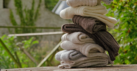 A tall stack of Merino wool socks seen in a rustic setting, resting on a piece of wood with a house in the background.
