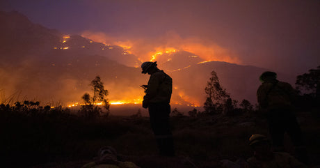Two silhouetted firefighters stand in front of a massive wildfire at night, with flames engulfing a smoky mountain range.