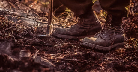 A close-up of rugged boots on uneven, debris-covered ground with a shovel partially dug into the soil nearby.