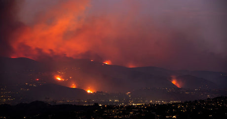 A wildfire burns across hills under a smoky, glowing orange sky, with a city’s lights visible in the dark foreground.
