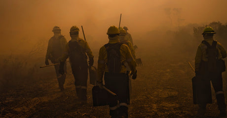 A group of wildland firefighters wearing yellow gear and hard hats walk through dense orange smoke on rough terrain.