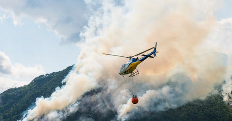 A firefighting helicopter with an orange water bucket flies through dense smoke over a wildfire in a forest.