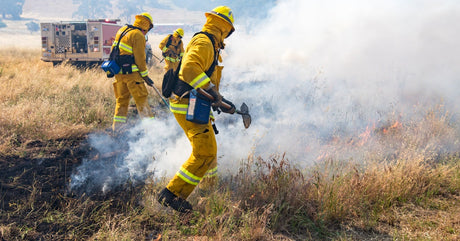 Firefighters in yellow gear cut a smoking grass fire with hand tools as a fire engine parks nearby in a dry field.