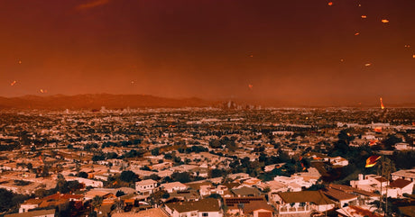 An orange smoke-filled sky hangs over Los Angeles as embers drift above houses and a distant skyline.  File Name